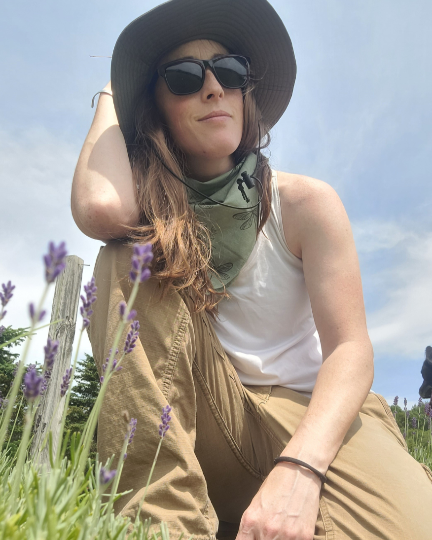 small business owner sitting in a field of lavender wearing sunglasses, a hat, and a scarf.